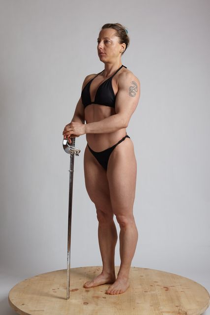 a woman stands in a studio with a grey background. she's wearing a black bikini and is holding a silver sword in her left hand. she has muscular arms and is looking to the right. she's standing on a wooden platform.