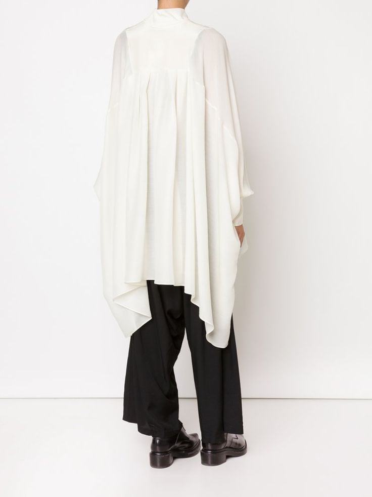 a woman is standing with her back to the camera wearing a white blouse with long, billowy sleeves and black pants with black shoes. the blouse is oversized and appears to be made of a sheer fabric. the pants are widelegged and slightly cropped. the shoes are black leather loafers. the background is a plain white wall.