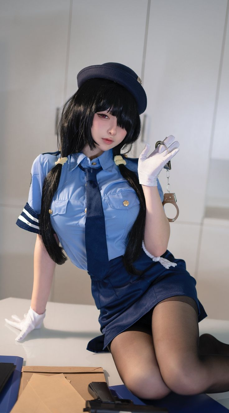 a woman is posing in a police uniform. she is wearing a blue hat, a light blue shirt with a dark blue tie, a dark blue skirt, and black stockings. she is holding handcuffs in her right hand and has white gloves on both hands. a gun and some evidence bags are on the table next to her.