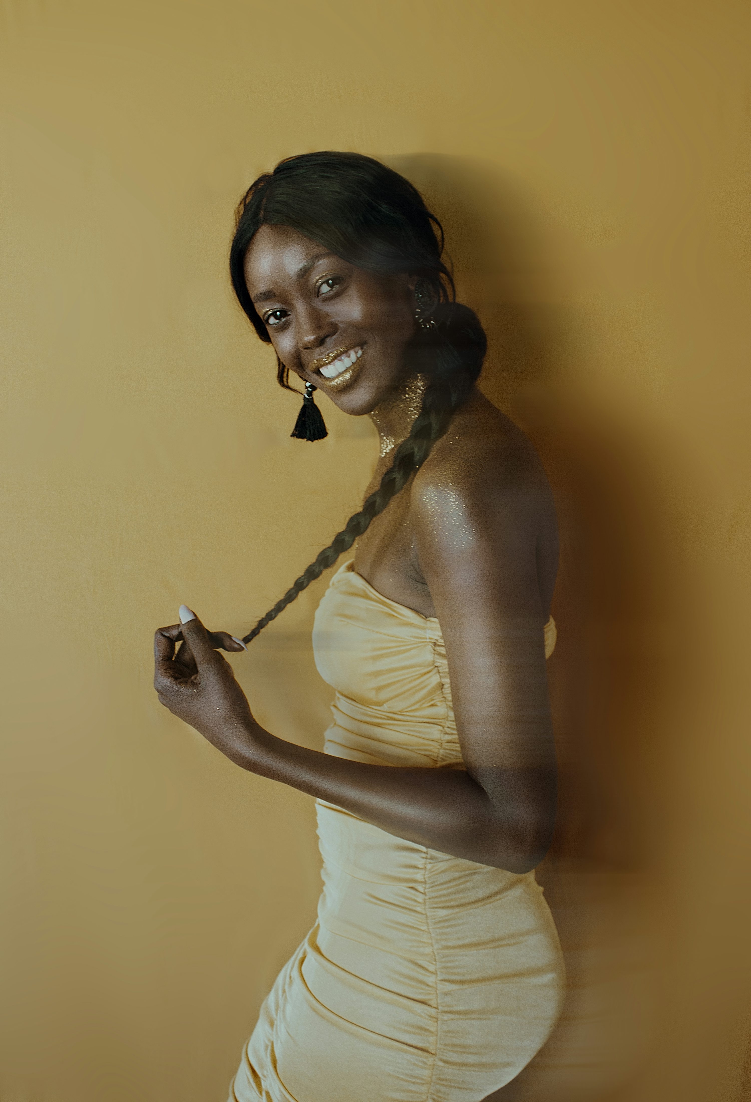a young black woman with a confident smile stands against a yellow background. she's wearing a gold dress with a ruched bodice and has gold glitter on her shoulders. her hair is styled in a long braid adorned with gold. she wears black tassel earrings. the overall mood is elegant and sophisticated.