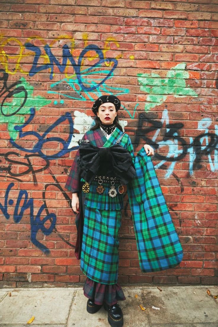 a woman stands in front of a brick wall covered in graffiti. she is wearing a plaid kimono, a black beret, and platform shoes. she is also wearing jewelry.