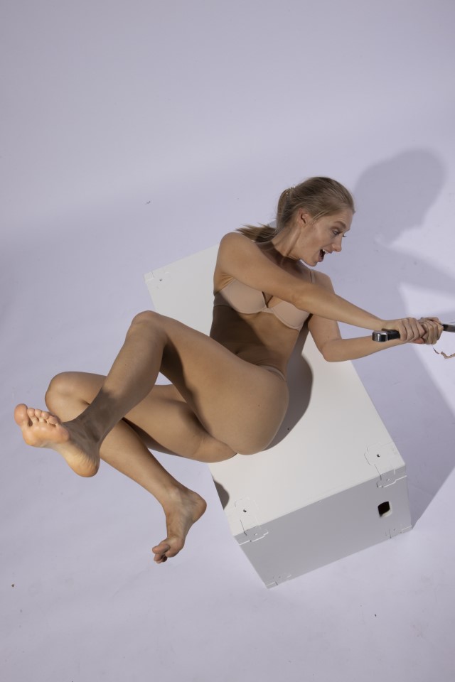a woman with blonde hair tied back is positioned on a white box in a studio setting with a light background. she is barefoot and wears a beige bra and body stocking. her expression conveys surprise or fear, and she is holding a tool in her hand. the woman's body, arms, and legs are visible.