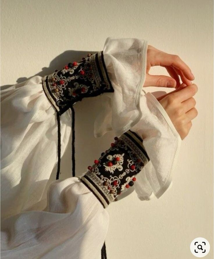 a closeup of a person wearing a white dress with black and red embroidered cuffs with beads. the dress is made of a light weight fabric and has a ruffled sleeve. the embroidered cuffs are intricate and have a floral design. the image is taken from a high angle, looking down at the arm of the person. the background is a light brown wall, which creates a simple and elegant aesthetic. the overall tone of the image is one of elegance and sophistication.