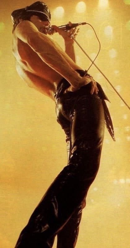 a man, wearing black leather pants, stands on a stage, holding a microphone in his hand. he is singing and looking towards the audience. he is illuminated by a spotlight, and the background is blurred. the image is most likely from a live concert. the performer is freddie mercury, lead singer of queen, posing in a photo shoot.