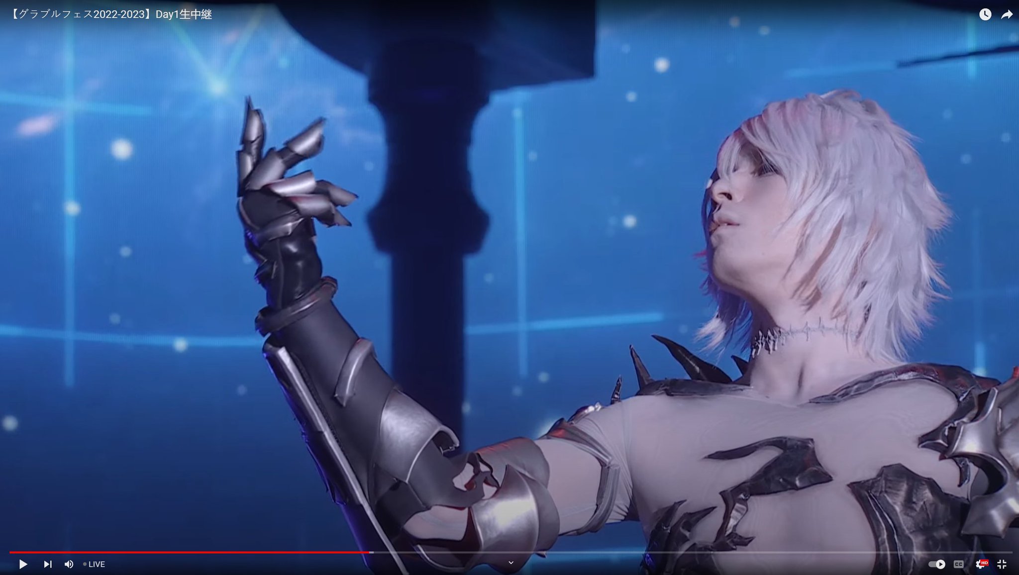 this is a photo of a woman cosplaying an anime character. she is wearing a silver and black armor with glowing details. her hair is white and she is looking up with a slight smile on her face. she is posing on stage in front of a blue background with lights and a blurred audience in the background.  she is looking up as if she is performing for an audience.