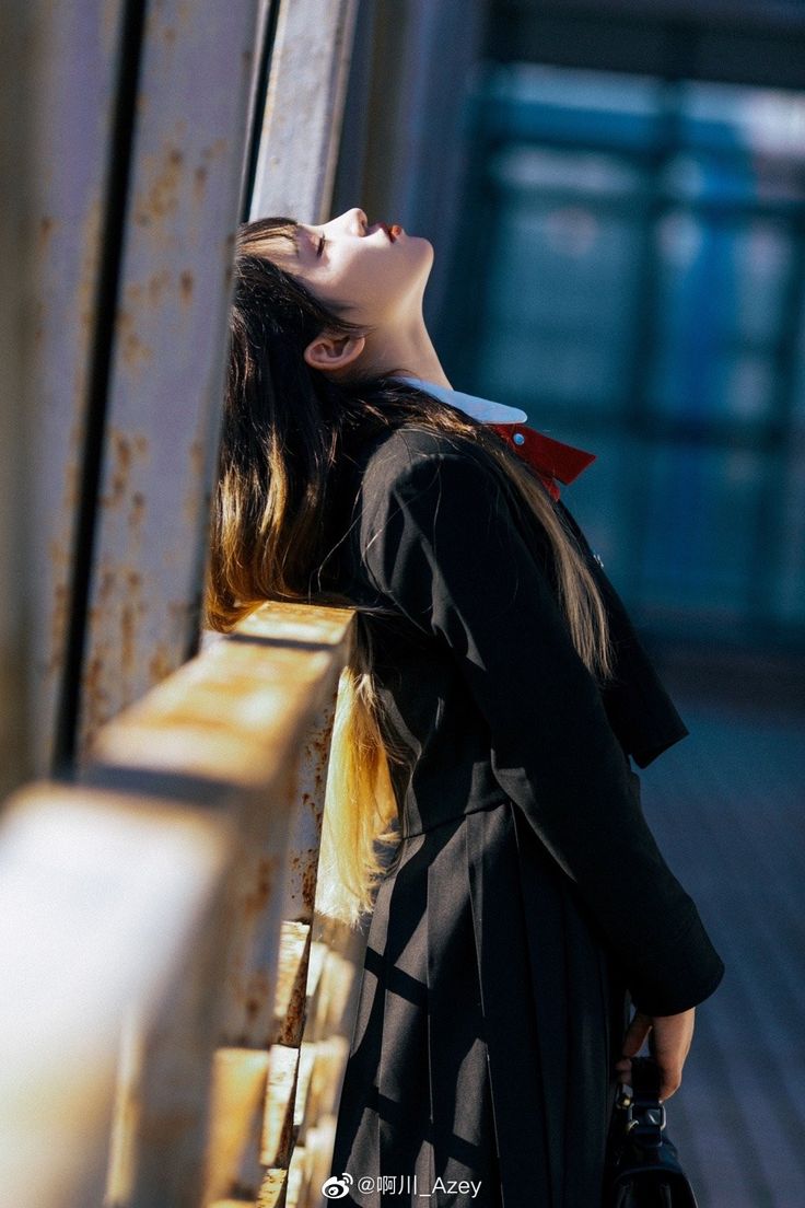 a young woman in a school uniform leans against a rusty bridge railing, her head tilted back as if looking up at the sky. she has dark hair with blonde highlights and wears a black jacket over a white shirt with a red collar. the scene is set outdoors, with a slightly blurred background of the bridge structure. the woman appears pensive or wistful.