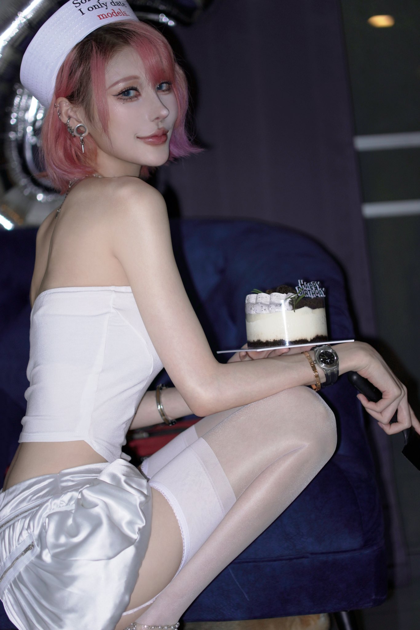 a young, attractive asian woman with short pink hair is sitting on a dark blue chair. she is wearing a white sailor hat, a white strapless top, and white stockings. she is holding a small birthday cake. the setting appears to be indoors at night.  the overall mood is elegant and glamorous.
