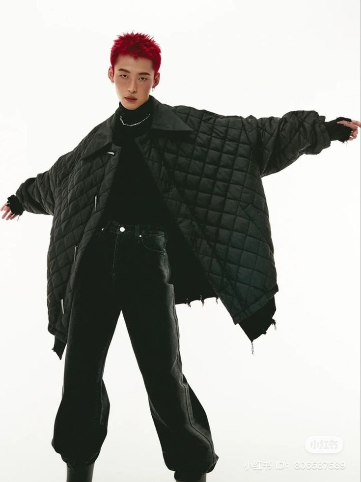 a man with red hair is wearing a black jacket and black pants. he is standing in a studio and posing for a photo. he is looking at the camera with a serious expression. he is wearing a stylish outfit.