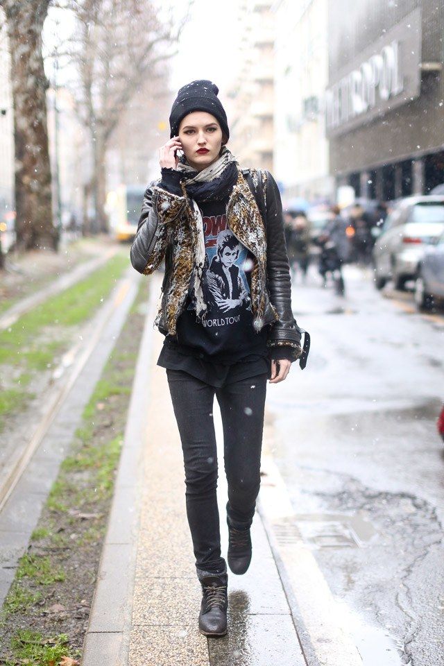 a stylish woman, possibly a model, walks down a city street on a cold, snowy day. she is talking on her phone and is dressed in a trendy winter outfit that includes a black beanie, a leopardprint jacket, a black scarf, black jeans, and black boots. her tshirt features a rock and roll band graphic.  the overall style is casual yet cool and urban. the background is blurred, showing a city street with buildings and other people.