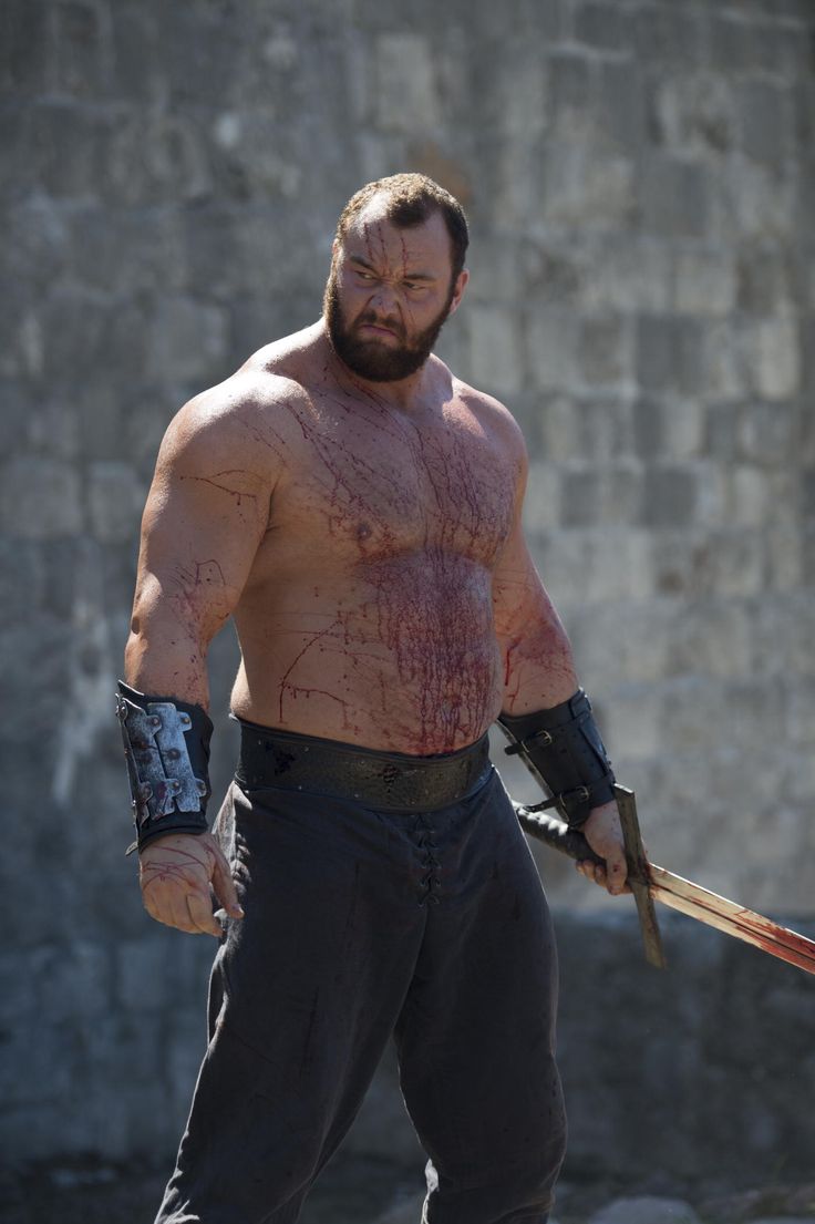 a large, muscular man in a dark, medieval costume and armor stands against a gray wall, holding a sword in his right hand, looking off to the side with a dramatic expression. he is covered in blood and looks wounded and intense.