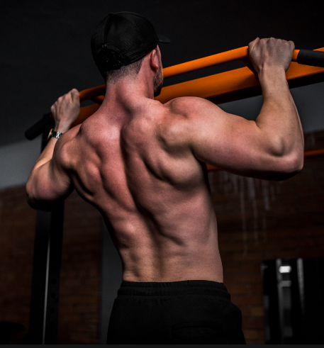 a muscular man is shown from the back, performing a pullup on an orange and black bar in a gym. the perspective is from a low angle, emphasizing the man's back muscles and the effort involved in the exercise. the lighting is artificial, typical of a gym setting.