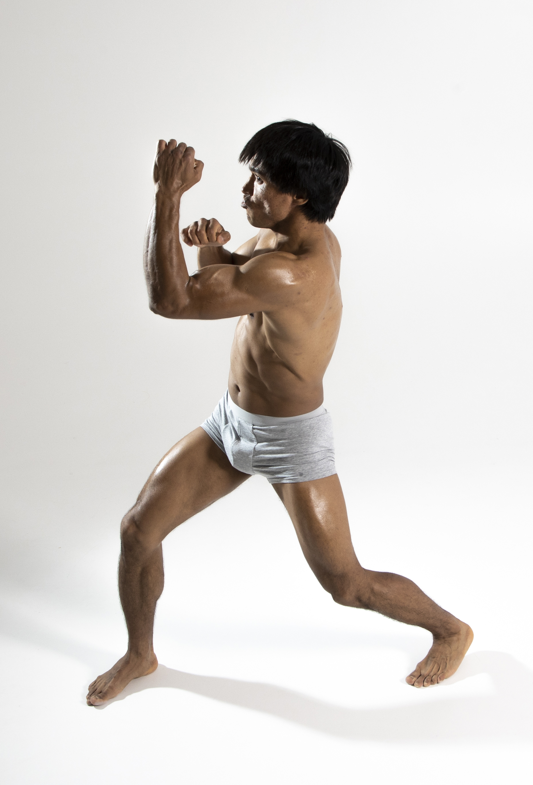 a shirtless man wearing gray shorts is captured in a boxing pose against a white background. he is muscular and sweaty, suggesting intense physical activity. his fists are raised as if ready to fight, and his body is angled in a classic boxer's stance. the overall image conveys athleticism, fitness, and strength.