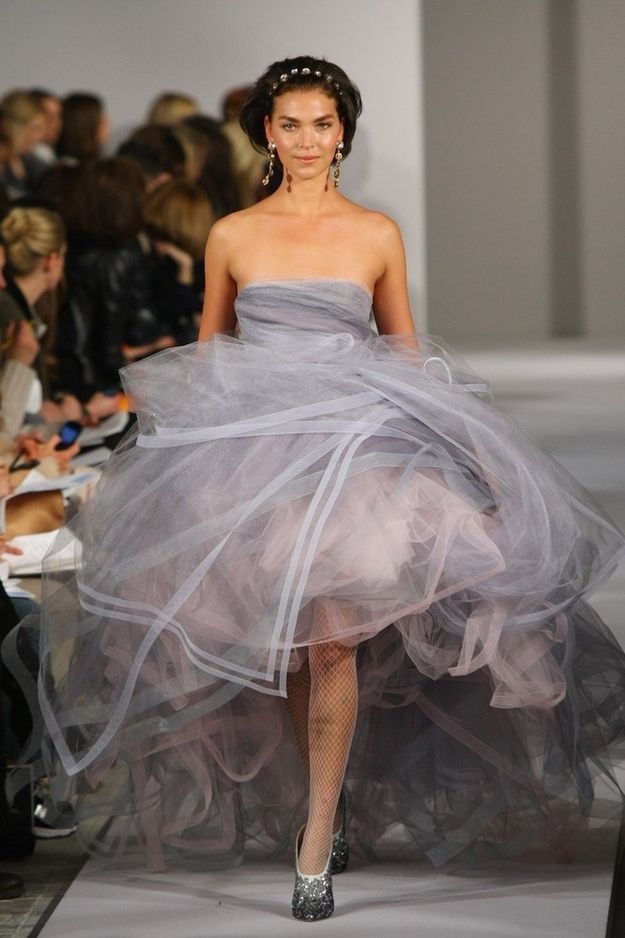 a model walks the runway in a layered tulle dress with pink and grey tones. the dress is cinched at the waist with a black belt, and the model is wearing silver, sparkly shoes. she also has her hair up in a bun, and is wearing makeup with a nude lip and a silver eyeshadow.