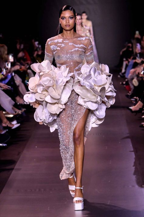 a fashion model walks down the runway wearing a white, sheer, sequined gown with a voluminous white ruffle skirt. the dress is designed with a plunging neckline and long sleeves. she wears white platform heels and her hair is styled in a sleek ponytail. the model is smiling and looking directly at the camera. the background is filled with audience members and other models.