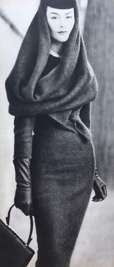 a black and white photo of a woman in vintage clothing posing for a fashion shoot. she is wearing a hat, a scarf, gloves, a purse, and a dress that looks very chic.