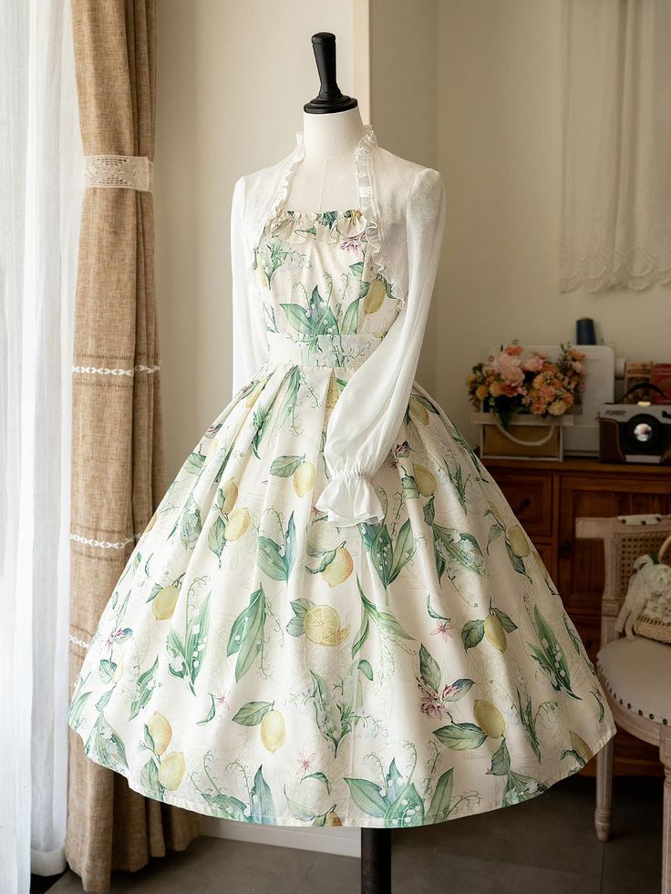 a white and green floral dress with lemon prints displayed on a mannequin. the dress is accompanied by a white shrug. the scene is indoors with natural lighting and some furniture.