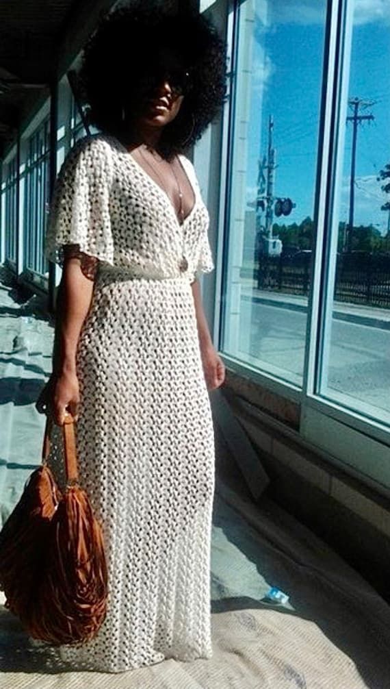 a young african american woman with a voluminous afro hairstyle wears a white crochet maxi dress and brown fringed handbag. she's standing outdoors near a large window of a building, sunlight illuminating the scene.  she's wearing sunglasses and appears fashionable and stylish.