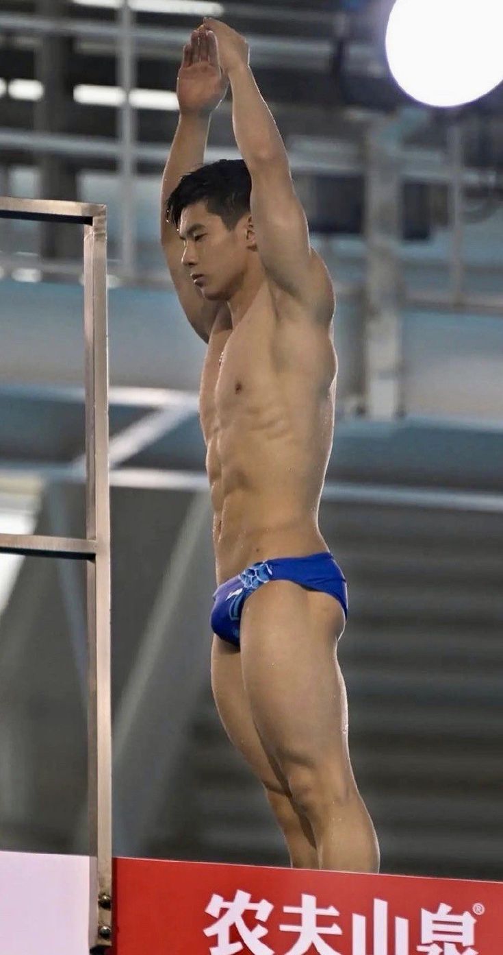 a young, muscular male diver stands on a diving platform, poised to begin his dive. he is wearing a blue pair of swimming trunks. his body is athletic and toned. the background is slightly blurred, indicating a competition setting. the red sign in the foreground displays chinese characters. the image focuses on the athlete's physique and readiness.