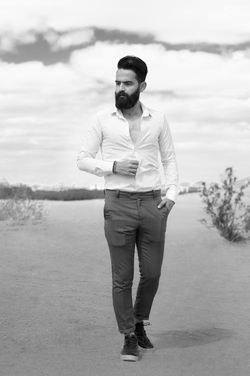 a black and white full shot of a stylish man with a beard and mustache standing on a road. he is wearing a white shirt, darkcolored pants and darkcolored sneakers. the man is looking down and slightly to his left. the background is a blurry landscape with a road and some vegetation. the sky is overcast.