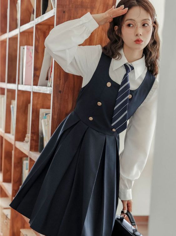 a young woman is posing in a preppystyle outfit. she's wearing a white shirt, a navy blue jumper dress with gold buttons and a pleated skirt, a striped blue and white tie, and round glasses. she's carrying a black bag and is standing in front of a wooden bookshelf filled with books. the lighting is bright and natural, and the image is taken from eye level.