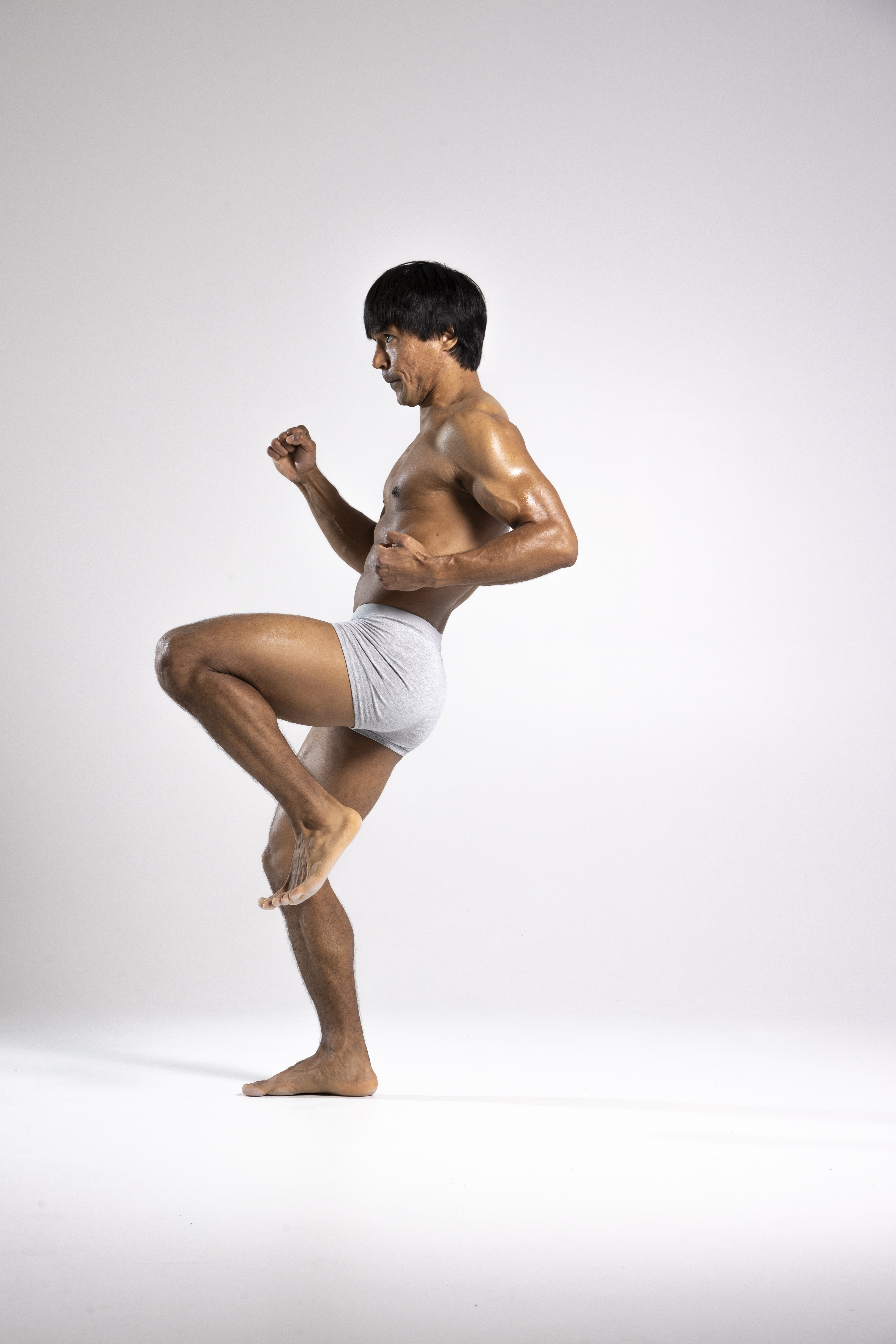 a man stands shirtless in boxer shorts, with his knee raised, fists clenched, and appearing to be in a martial arts pose.
