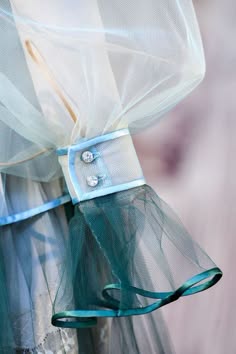 a closeup shot of a delicate blue tulle dress with a large ruffle at the sleeve. the dress is adorned with a ribbon and embellished with small silver buttons. the fabric is sheer and flowing, giving the dress a light and whimsical feel.