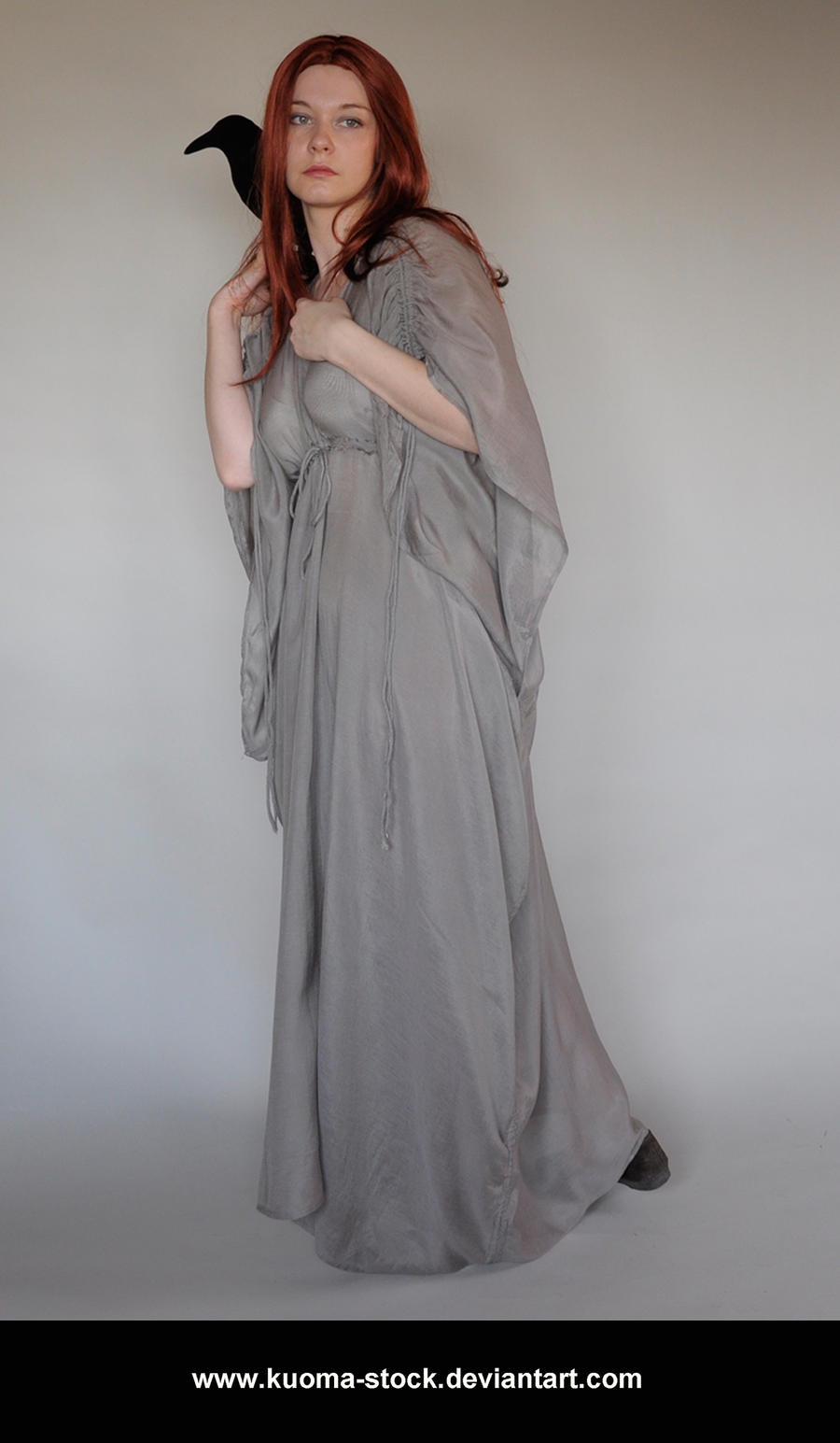 a fullbody shot of a young redhead woman, wearing a long, flowing, grey medievalstyle dress, posing in a studio against a plain light background. she has long, loose red hair and is holding a black raven or crow on her shoulder. her expression is somewhat mysterious and serious. the image has a fairytale or fantasy feel.  the watermark "www.kuomastock.deviantart.com" is visible at the bottom.