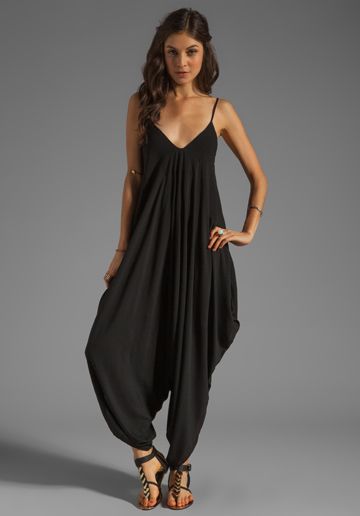 a woman is standing in a studio wearing a black jumpsuit with a vneck and sandals. she is posing for a fulllength shot.