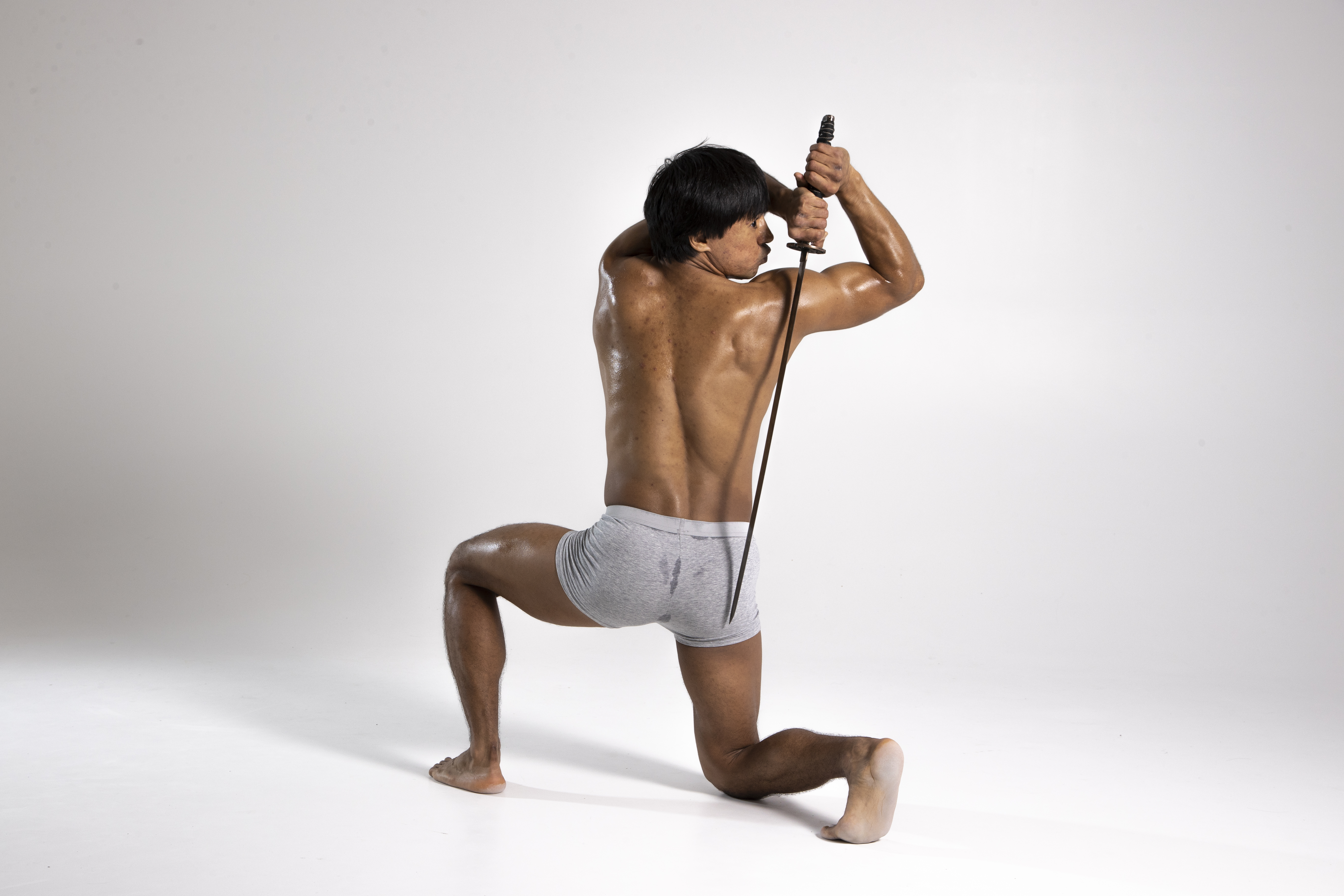 a shirtless man in gray underwear is kneeling with one knee on the ground, holding a sword behind his head.