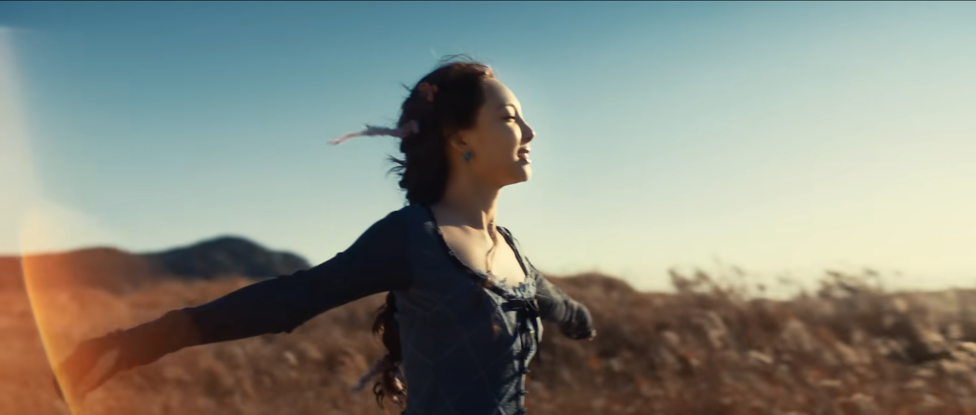 a woman in a blue dress is standing in a field with her arms outstretched. the sun is setting behind her, casting a warm golden glow on the scene. she has a feather in her hair.