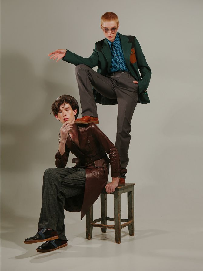 two models are posing in a studio setting. the woman is standing with one leg on a stool while the man is sitting on the stool. they are both wearing stylish outfits including a brown coat, a green blazer, and grey pants.  the woman is wearing a patterned shirt, and the man is wearing a brown hat. they both have their hair styled in a modern way and are looking at the camera with confident expressions.