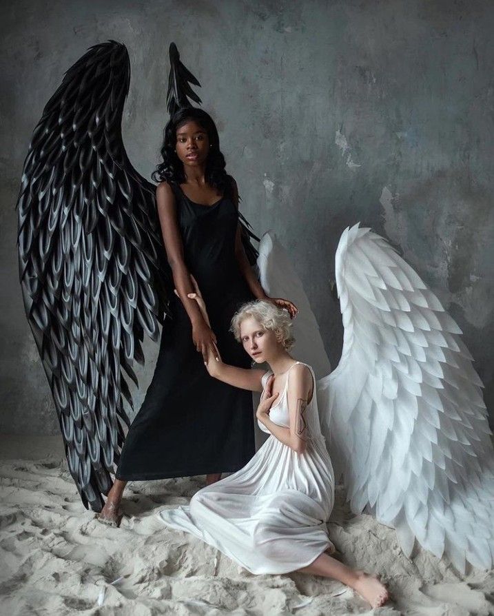 two women, one with black angel wings and the other with white angel wings, are standing in a studio setting. the woman with the black wings is wearing a black dress and has dark skin. the woman with the white wings is wearing a white dress and has fair skin. they are both looking at the camera with serious expressions on their faces. the lighting in the image is dramatic and creates a strong contrast between the two women and their wings. the image has a mystical and spiritual feel.