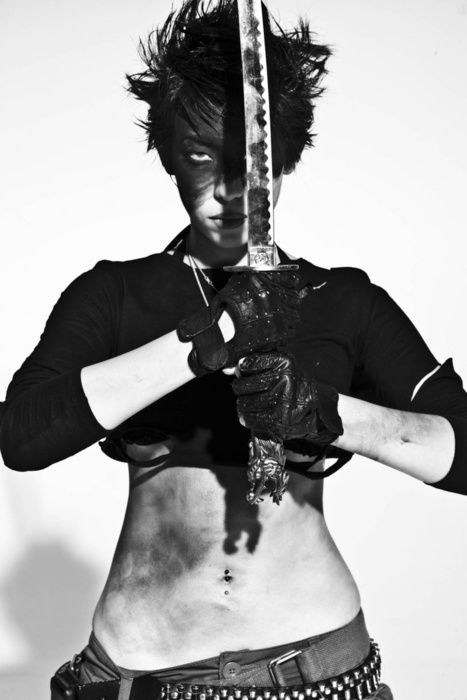 a woman wearing a black outfit poses while holding a katana sword. she's wearing a black top and pants, with gloves on her hands, and a belly piercing. her hair is styled up.