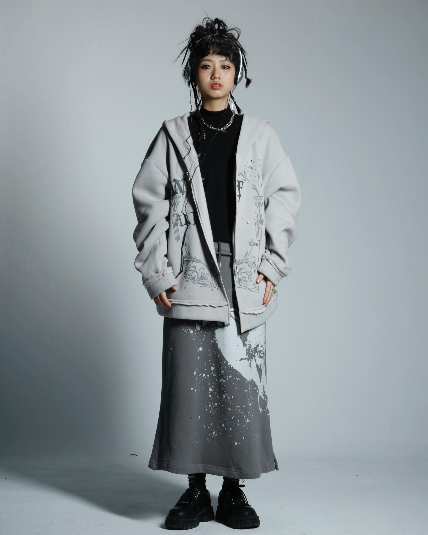 the image shows a young woman standing against a light grey background. she's wearing a light grey oversized hoodie with a graphic design and a dark grey long skirt with a splatterpaint and graphic design. her hairstyle features space buns and she has on black headphones around her neck.  she is wearing black shoes and a black top under the hoodie. the overall style is casual yet edgy, incorporating elements of streetwear and alternative fashion. the pose is relaxed and natural, with a slight tilt of the head.  the image appears to be a professional fashion photograph.