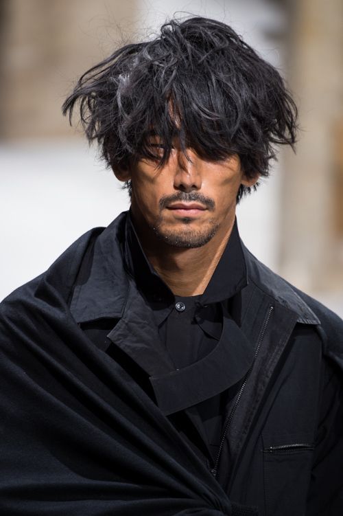 a closeup portrait of a male model with messy black hair covering his forehead and eyes. he has a short beard and mustache and is wearing a black jacket with a large collar that drapes over his shoulders. the background is blurred, suggesting a runway setting.