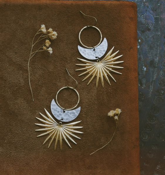 a pair of gold and silver sunburst earrings are displayed on a brown suede background. the earrings feature a hammered silver halfmoon shape above a gold sunburst design. two small dried flowers are placed near the earrings.