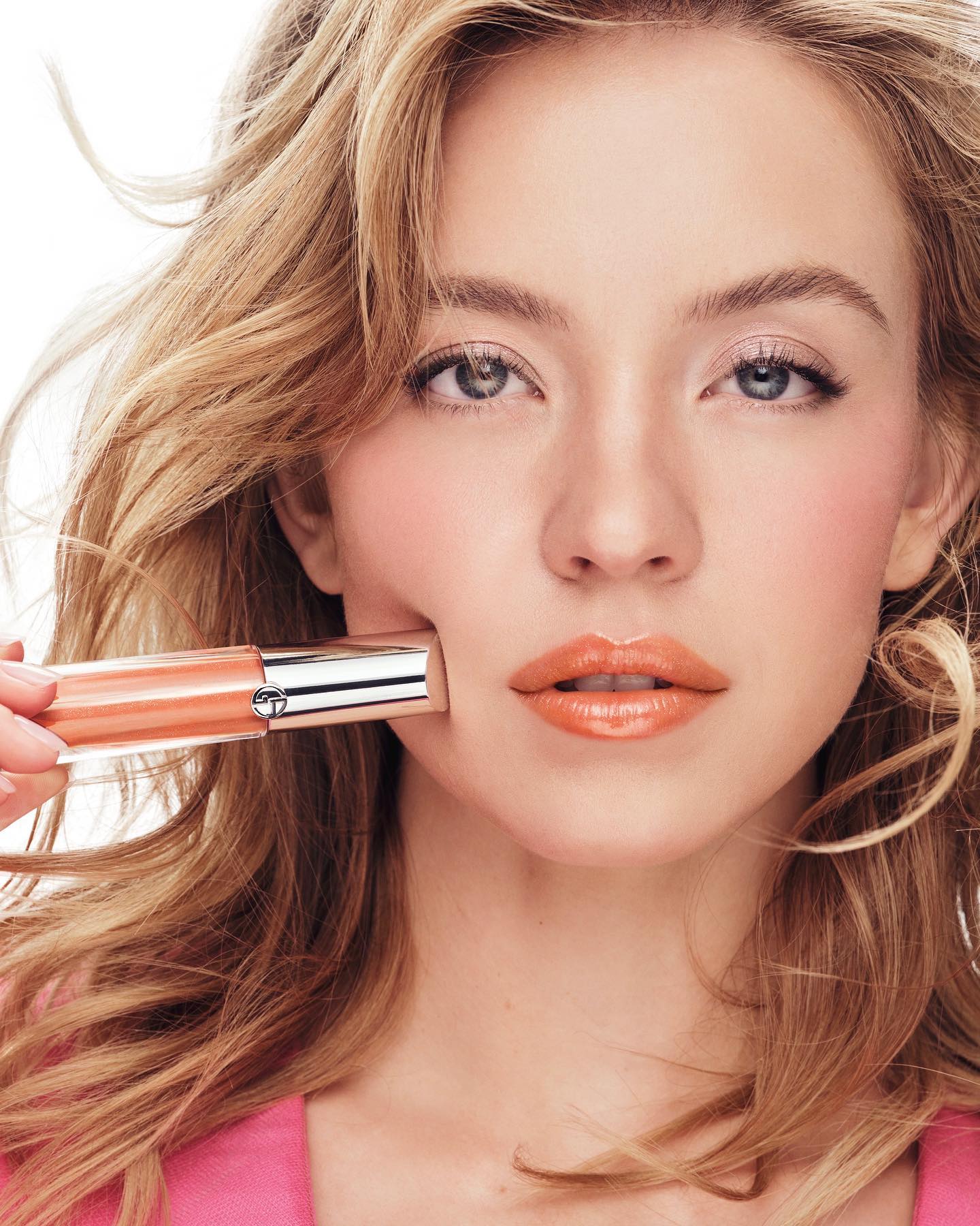 a closeup portrait of a young, blonde woman with long, wavy hair. she is holding a tube of peachtoned lip gloss or lipstick near her face. the woman is wearing minimal makeup, highlighting her natural beauty.  the lipstick's color is a soft, peachy coral. she is wearing a pink top. the image is likely an advertisement for the beauty product. the style is high fashion and glamorous. the background is pure white, drawing focus entirely to the model and product.