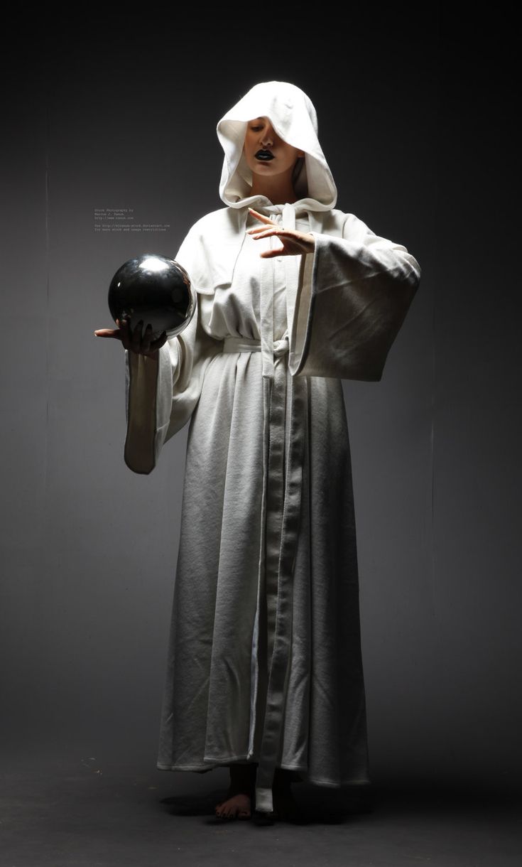 a woman in a white hooded robe holds a crystal ball in front of her. she has dark eye makeup and a serious expression. the image is dark and atmospheric, creating a mysterious and mystical mood.