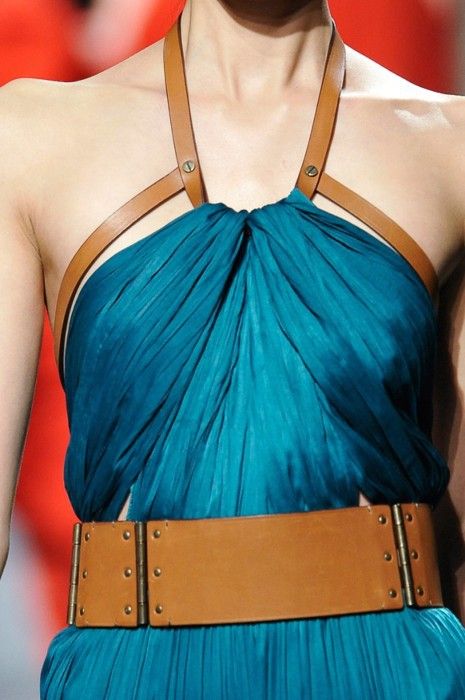 a closeup shot of a woman wearing a blue pleated dress with brown leather straps forming a halter top and a wide brown leather belt around the waist. the dress has a draped and gathered design at the neckline. the belt has metal accents and a rigid rectangular shape. the background is a blurred red.
