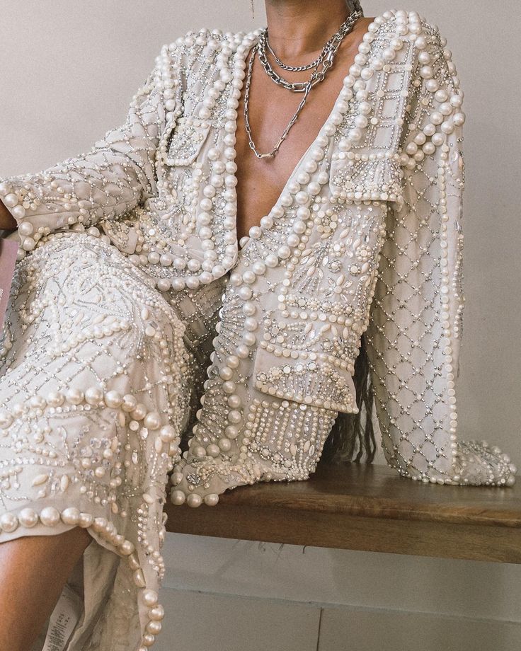 loseup shot of a woman modeling a luxurious, pearlembellished jacket and dress. the outfit is predominantly offwhite or beige, heavily adorned with pearls and intricate beading. the jacket features a lapel collar and a slightly open front, showcasing several delicate necklaces layered around the model's neck. the dress underneath seems to match the jacket in design and embellishments. the model’s dark hair is visible along the side of the jacket, and a wooden surface is subtly visible in the background. the overall aesthetic is elegant, luxurious, and possibly bridal or highfashion related.