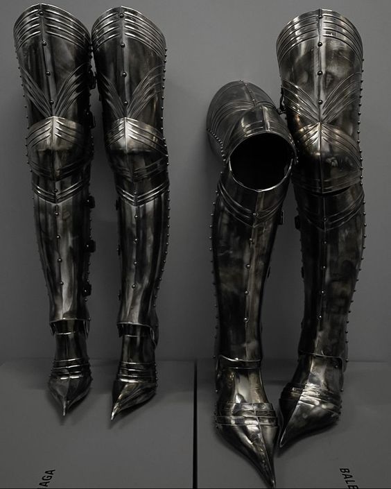the image showcases a pair of intricately crafted medieval leg armor, known as greaves, displayed against a neutral gray background. the greaves are made of polished steel, reflecting the artificial light of the museum environment. the left greave is fully assembled, while the right one is shown disassembled, offering a view of its inner structure. the pointed toes and articulated joints highlight the craftsmanship and functionality of these pieces designed for both protection and mobility in battle.