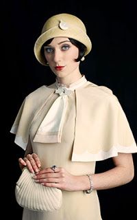 a woman in a 1920s flapper outfit. she is wearing a cloche hat, a white dress with a cape, white gloves, and a pearl necklace. she is holding a white purse and a small ring with a large blue stone.
