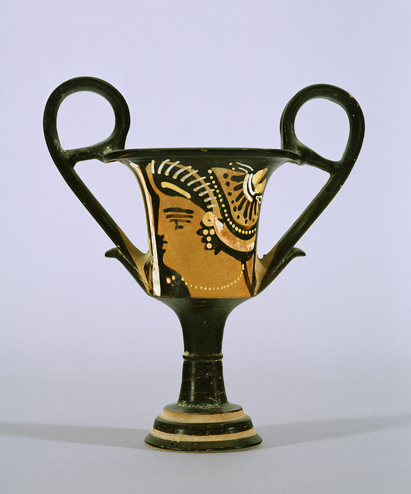 a blackfigure kylix with a stylized design.  the kylix is a twohandled drinking cup, and the design is a stylized figure with wings and a head. the body is black, with a band of orange in the center, and the handles and base are black.  it is typical of pottery from the archaic and classical periods in greece.