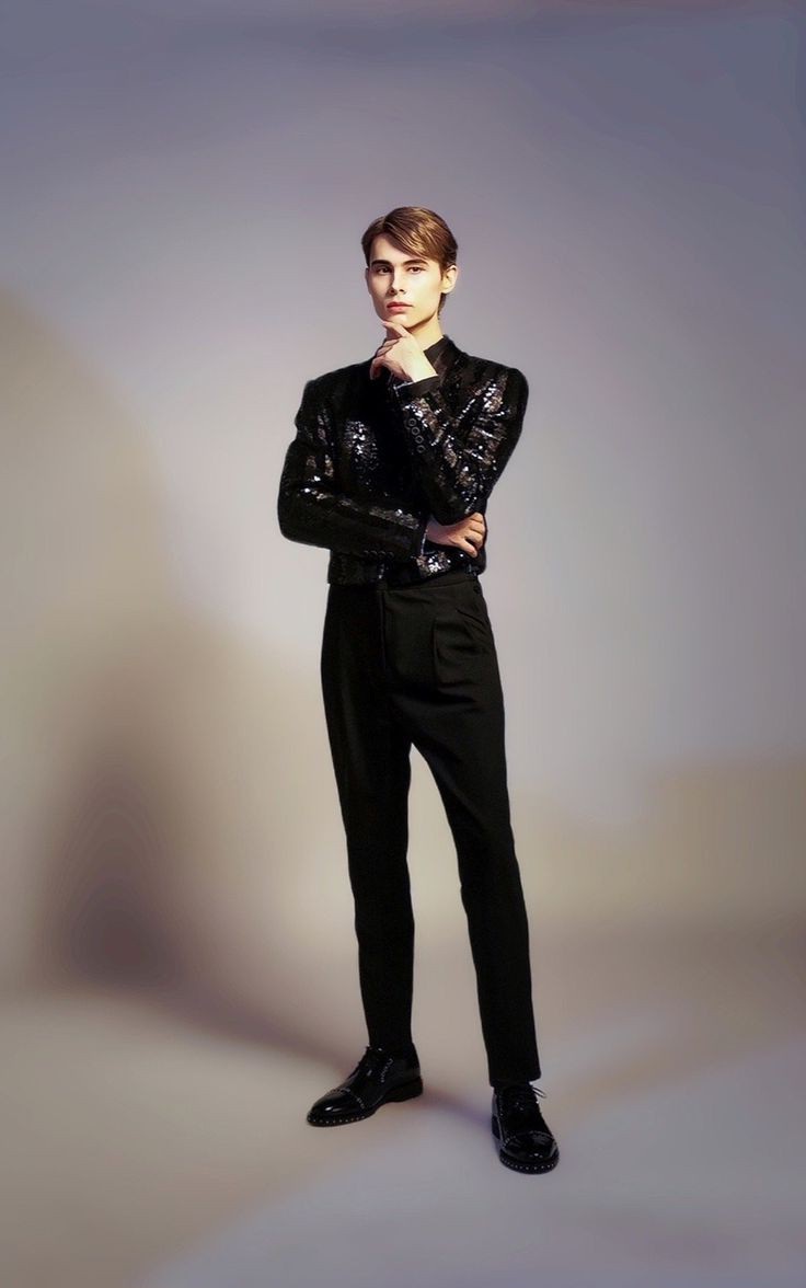 a fullbody shot of a young male model posing against a light purple backdrop. he's dressed in a stylish black sequin jacket, black trousers, and black leather shoes. his posture is confident, and he has his hand on his chin. the overall style is elegant and highfashion.  the image is welllit, creating a professional and sophisticated feel.