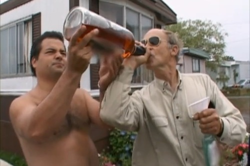 a shirtless man holds a very large bottle of liquor for an older man wearing sunglasses and a buttondown shirt, who is drinking from it. they are outside in what appears to be a trailer park. the older man is also holding a white cone and a bottle.