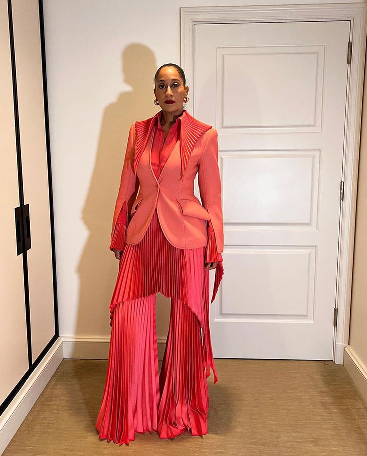 a woman is standing in a hallway wearing a red and pink pleated suit. the suit consists of a blazer, trousers, and a shirt. the blazer is pink and has a single button closure. the trousers are red and have a wide leg. the shirt is red and is tucked into the trousers. the woman is wearing earrings and lipstick. the background is a hallway with a door and a shadow on the wall.