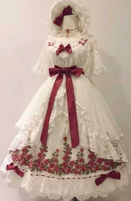 a white lolita fashion dress with red rose embroidery is displayed on a mannequin. the dress is adorned with lace trim, a red ribbon belt, and a matching bonnet. the dress falls to the floor with multiple layers of fabric and lace.