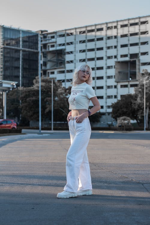 a blonde woman is standing in a city street, posing for a photo. she's wearing a white crop top, white pants, and white shoes. the background is a large, modern building. the photo has a casual and stylish vibe.