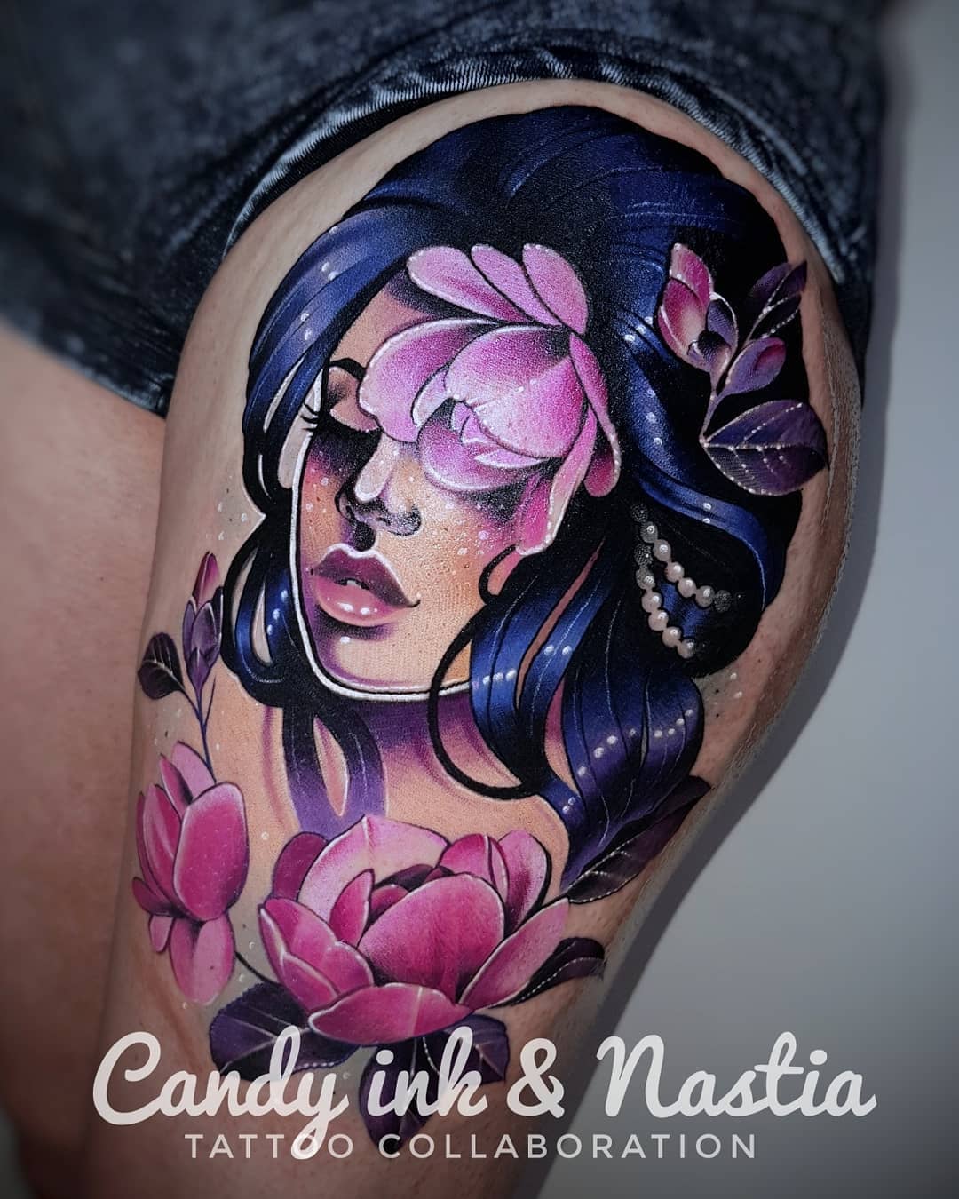 a woman with blue hair and pink flowers tattooed on someone's upper thigh. the woman has one eye closed and is looking downward. there are two large magnolias, one partially covering her face and the other below her chin, and a few smaller flower buds around the larger flowers. the woman is wearing small pearl earrings. the tattoo has a soft, painterly style and uses shades of pink, purple, and blue. the tattoo artists' names, "candy ink" and "nastia," are written in cursive script below the tattoo along with the phrase "tattoo collaboration."