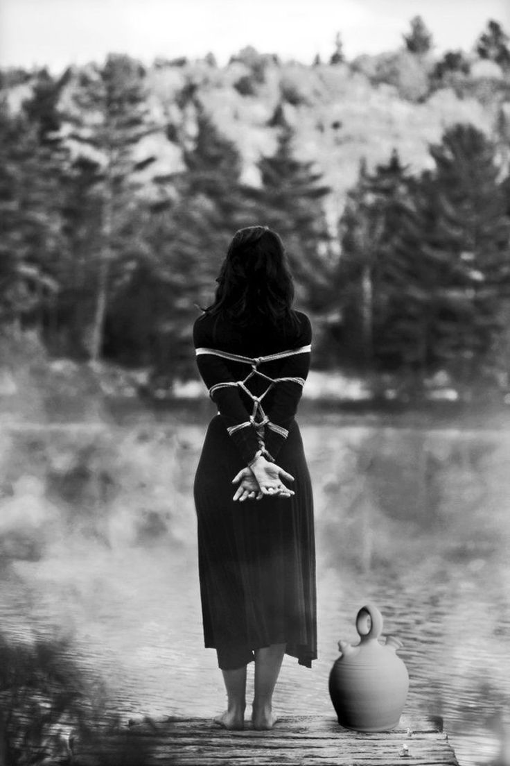 a black and white photograph depicts a woman standing with her back to the camera, tied up with rope around her wrists and torso. she is wearing a long black dress and is standing on a wooden dock by a lake in a wooded area. a clay pot sits on the dock beside her. the image is moody and atmospheric, creating a sense of mystery and contemplation.  the overall feeling is one of isolation and possibly captivity.